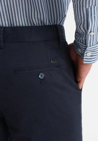 Pantaloni blu navy con una tasca sul retro chiusa da bottone, caratterizzati da una finitura leggermente strutturata e un piccolo logo metallico.