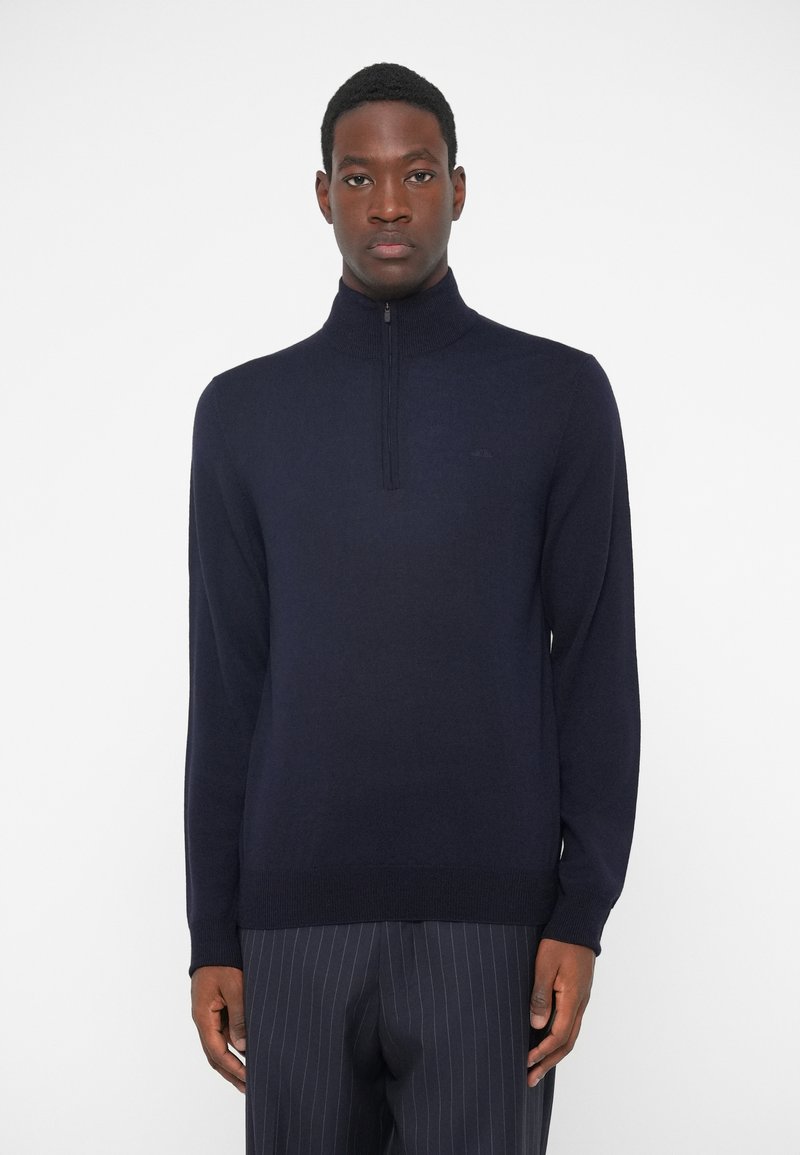Granatowy sweter z dzianiny z wysokim kołnierzem, wyposażony w zamek błyskawiczny na ćwierć długości. Projekt jest dopasowany, posiada długie rękawy oraz ściągacze u dołu i na mankietach.