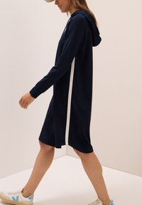 Merellinen hupparimekko valkoisilla sivuraidoilla, valmistettu pehmeästä kankaasta. Ominaisuuksina pitkät hihat ja hieman löysähkö mitoitus, päättyen polven ylle.