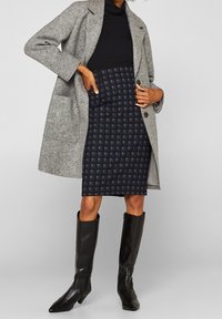 Grå ullkappa över en svart polotröja, kombinerad med en marinblå rutad pennkjol och knähöga svarta läderstövlar.