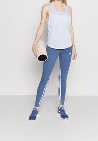 Top de tirantes azul claro combinado con leggings azul oscuro que tienen rayas laterales blancas, sostenido con un rodillo de espuma beige texturizado. Zapatos deportivos oscuros.