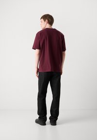 Bordeauxfarbener Baumwoll-T-Shirt mit relaxed Fit, kurzen Ärmeln und Rundhalsausschnitt, kombiniert mit schwarzen Jeanshosen und schwarzen Schuhen.