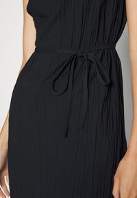 Robe noire texturée cintrée à la taille avec une fine ceinture nouée, laissant apparaître une partie des bras et du torse de la personne qui la porte.