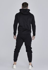 Čierny set mikiny a teplákov, s jemnou textúrou, slim fit a rebrovanými manžetami. Oblečenie má kapucňu a žiadne viditeľné logá ani vzory.