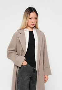 Palton bej din lână, cu o croială lejeră, revere mari și mâneci lungi, purtat peste un top negru și o cămașă deschisă la culoare, cu buton.