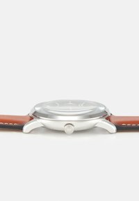 Runder Silberuhrgehäuse mit klarem Glaszifferblatt. Braunem Lederarmband mit kontrastierenden Nähten. Seitliche Krone zur Einstellung sichtbar.