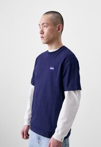 Tricou din bumbac bleu marine, suprapus peste o cămașă albă cu mâneci lungi, având un logo mic, gât rotund și o croială lejeră.