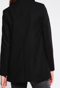 Blazer de lana negra con diseño estructurado, abertura trasera única, mangas largas y textura suave. Silueta simple sin elementos metálicos visibles.