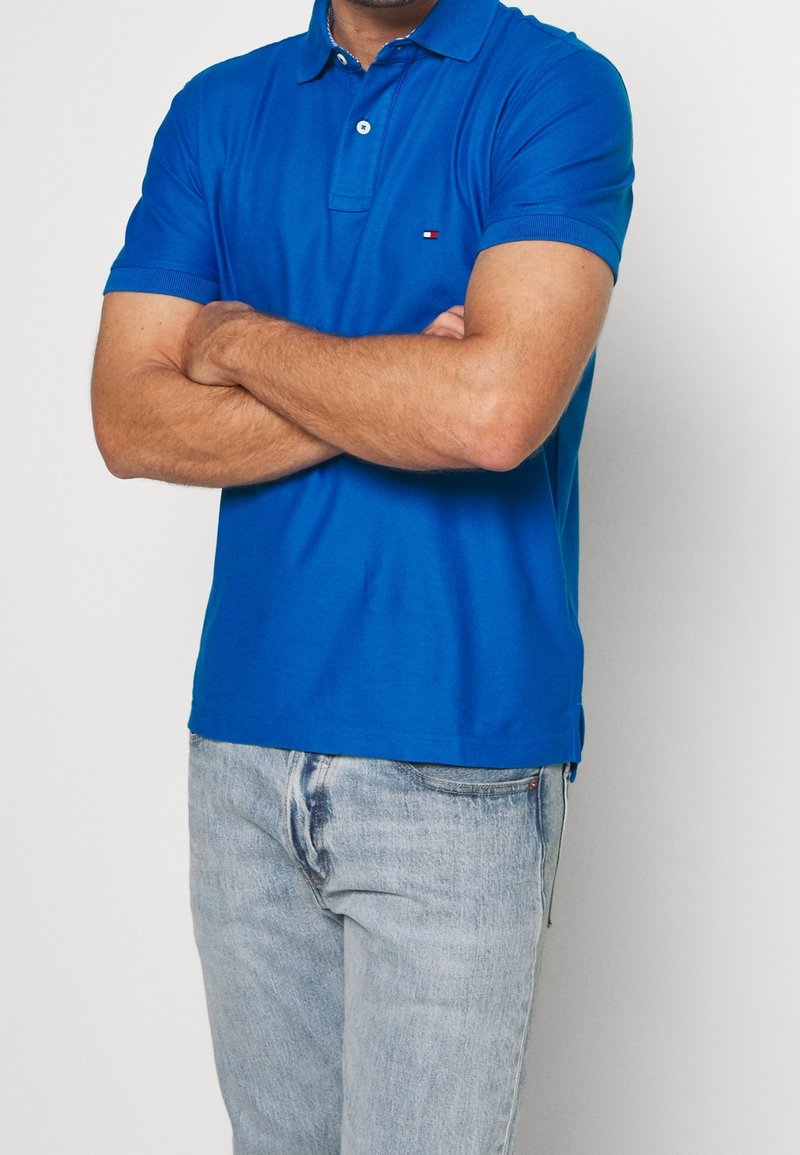 Homme portant un polo bleu à manches courtes, les bras croisés et un jean bleu clair délavé sur un fond uni.