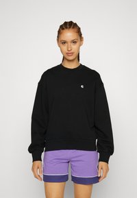 Schwarzer Pullover mit gerippten Bündchen und Saum, versehen mit einem kleinen weißen Logo auf der Brust. Getragen mit lila Shorts mit kontrastierenden Nähten.