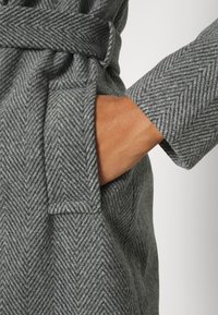 Mano nella tasca di un cappotto di lana grigio a spina di pesce con cintura legata in vita, che mostra la texture del tessuto e un dettaglio ravvicinato della manica.