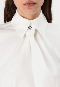 Hvit bluse med en strukturert krage, med en knytedetalj ved halsen og svarte broderte detaljer ved kragefoten.