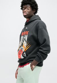 Μαύρο φούτερ με κουκούλα με ζωντανό πορτοκαλί και κόκκινο γραφικό, με θέμα το μπάσκετ. Φορεμένο πάνω από κόκκινο πουκάμισο, συνδυασμένο με ανοιχτό πράσινο τζόγκερ.