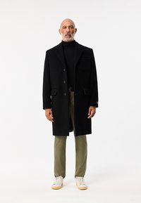 Zwarte wollen rok overjas met knopen, gestructureerde revers en een borstzak, gecombineerd met olijfgroene broek en witte sneakers.