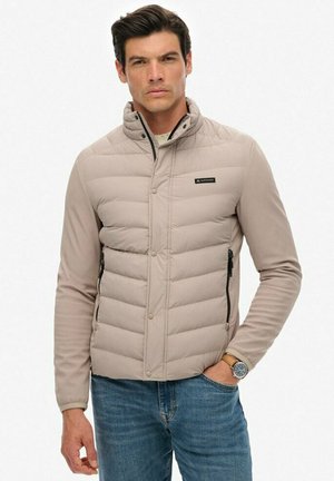 Beige pufferjack met een hoge kraag, horizontaal gestikt ontwerp, zijzakken met rits en een logo-opdruk op de borst. Draagt over een licht shirt.