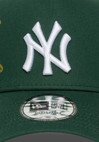 Casquette de baseball verte avec un logo "NY" brodé en blanc. Comprend un autocollant circulaire "New Era" étiqueté "Ajustable". Texture de tissu lisse.
