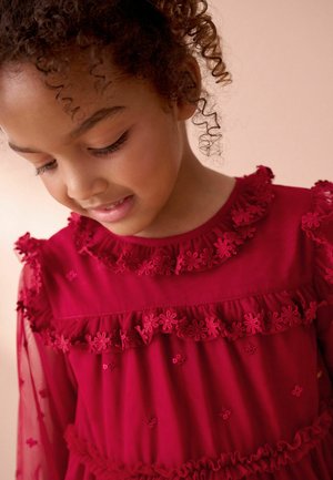 Ragazza giovane con capelli ricci guarda in basso, indossando un vestito rosso decorato con pizzo floreale e maniche trasparenti.
