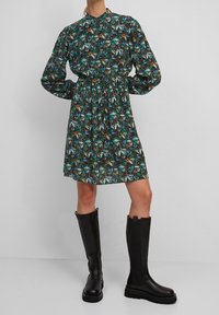 Vestido floral con una base oscura y patrones multicolores, mangas largas, cintura ceñida y un cuello alto. Combinado con botas negras hasta la rodilla.