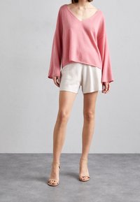 Rosa, locker sitzender Pullover mit V-Ausschnitt und langen Ärmeln, kombiniert mit hellbeigen Shorts. Der Stoff erscheint weich und hat eine entspannte Silhouette.