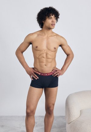 Jauns vīrietis ar cirtainiem matiem, tērpies melnās Tommy Hilfiger boksera apakšveļas biksēs, stāv ar rokām uz gurniem blakus baltai teksturētai atpūtas krēslam.