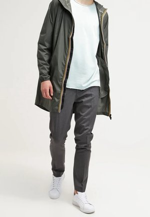 Mężczyzna idący w ciemnozielonym, ocieplanym płaszczu przeciwdeszczowym z kapturem, jasnoniebieskiej koszulce, szarych spodniach i białych trampkach na jasnym tle.