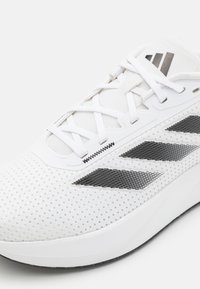 Scarpa sportiva bianca con tomaia in rete perforata, design a tre strisce nere e lacci piatti. Presenta un'intersuola bianca imbottita e una suola testurizzata.