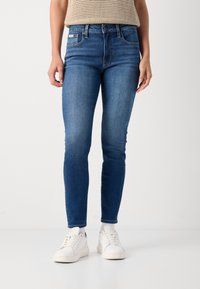 Modré úzke džínsy z denimu, s klasickým dizajnom piatich vreciek, stredne vysokým pásom a jemným vyblednutím, skombinované s bielymi teniskami.