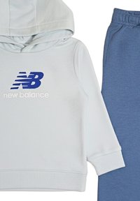 Felpa con cappuccio grigio chiaro con logo "NB" blu e testo "new balance", abbinata a pantaloni della tuta blu. Texture liscia, design casual.
