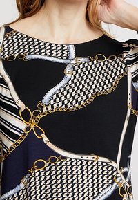 Svart topp med ett geometriskt bälte och kedjemönster i blått, vitt och guld. Korta ärmar med en kontrasterande randig design vid axeln.