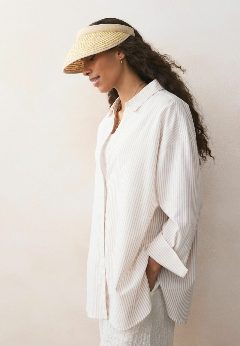 Bijela košulja s tankim okomitim prugama. Svijetla slamnata kapa s širokim obodom. Kovrčava kosa koja pada preko ramena.