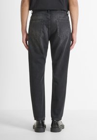 Jeans de denim negro con corte recto, con dos bolsillos traseros y costuras simples. Llevados con zapatos negros sobre un fondo neutro.