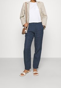 Blazer beige rayé sur un t-shirt blanc, associé à un pantalon en lin bleu marine et des sandales à glissière beige clair, accessoirisé avec un petit sac à main marron.