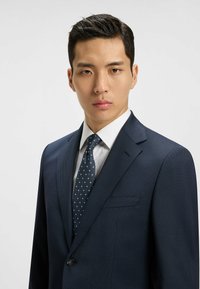 Marineblaues Nadelstreifen-Sakko mit Reverskragen, ausgestattet mit einem Ein-Knopf-Verschluss und einer Brusttasche, kombiniert mit einem weißen Hemd und einer Pünktchenkrawatte.