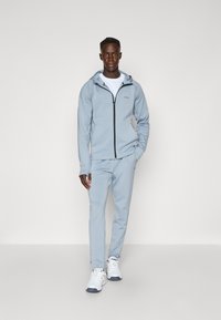 Ensemble de sport bleu clair en matériau doux, comprenant un sweat à capuche zippé et un pantalon assorti, avec des accents blancs et un détail de logo.