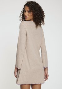 Vestido beige con mangas largas, corte holgado y dobladillo con volantes en tonos púrpuras. Textura suave y diseño minimalista.