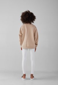 Beige överdimensionerad sweatshirt med ribbade muddar och fåll, kombinerad med vita leggings och ljusfärgade slip-on skor, sedd bakifrån.