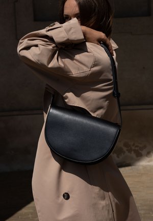 Femme en trench-coat beige portant un sac bandoulière en cuir noir en forme de demi-lune sur l'épaule, couvrant partiellement son visage avec le bras.
