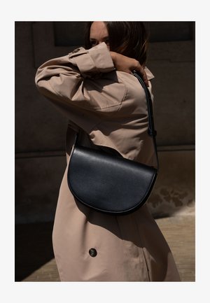 Femme en trench-coat beige portant un sac bandoulière en cuir noir en forme de demi-lune sur l'épaule, couvrant partiellement son visage avec le bras.