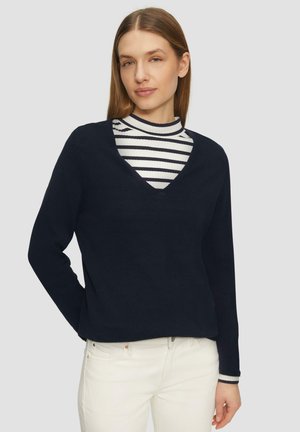 Marineblauwe trui met een V-hals en lange mouwen, gelaagd over een wit met marineblauw gestreepte coltrui. Zachte textuur en relaxte pasvorm.