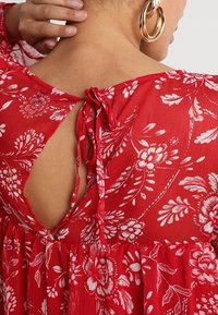 Robe florale rouge en tissu doux et texturé, avec un dos V, fermeture à nouer et motifs floraux blancs sur l'ensemble. Ourlet accentué.