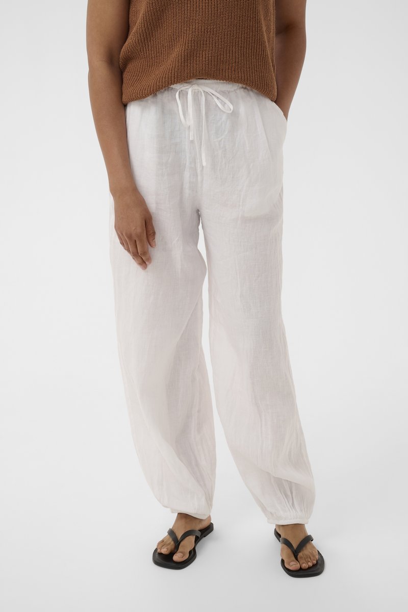 Personne portant un pantalon blanc ample avec cordon, un top marron sans manches en maille et des sandales noires à brides, debout devant un fond uni.