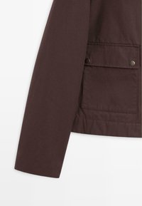 Braune Stoffjacke im Detail, die einen langen Ärmel und eine aufgesetzte Tasche mit Druckknopfverschluss zeigt; glatte Textur und lässiges Design.