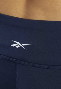 Granatowe legginsy do treningu mają gładki, elastyczny materiał z przeszywaną szeroką gumką w pasie i białym logo marki. Zbliżenie na górną część.