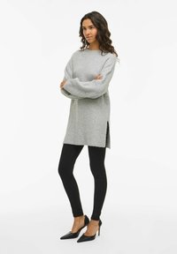 Sieviete ar čirkainiem matiem stāv ar roku salocītām, ģērbusies pelēkā lielizmēra džemperī, melnās legingās un melnās augstpapēžu kurpēs.