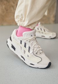 Weiße Sneakers mit schwarzen Akzenten und drei dunklen Streifen, ausgestattet mit einem glatten Obermaterial, strukturierten Details und einer dicken weißen Sohle. Rosa Socken sichtbar.