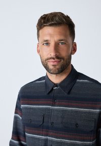 Camicia con bottoni caratterizzata da una base blu navy con strisce orizzontali in rosso, bianco e grigio. Ha un colletto e due tasche sul petto.