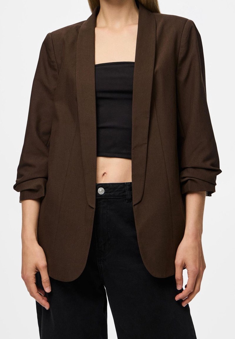 Femme portant un blazer marron aux manches retroussées sur un top tube noir et un pantalon noir, montrant le ventre découvert.
