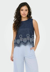 Top senza maniche in denim con ricami floreali bianchi lungo l'orlo, caratterizzato da un collo rotondo e una vestibilità comoda. Accoppiato con pantaloni di colore azzurro chiaro.