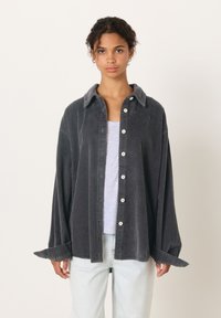 Moteris su garbanotais plaukais, dėvinti laisvą tamsiai pilką kordurovinę marškinių tipo palaidinę ant balto viršaus ir šviesiai mėlynas džinsus, stovinti priešais paprastą foną.