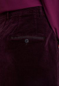 Pantaloni in velluto a coste borgogna con una tasca posteriore dotata di un bottone nero e una fascia orizzontale. Texture liscia con un distintivo motivo a coste.